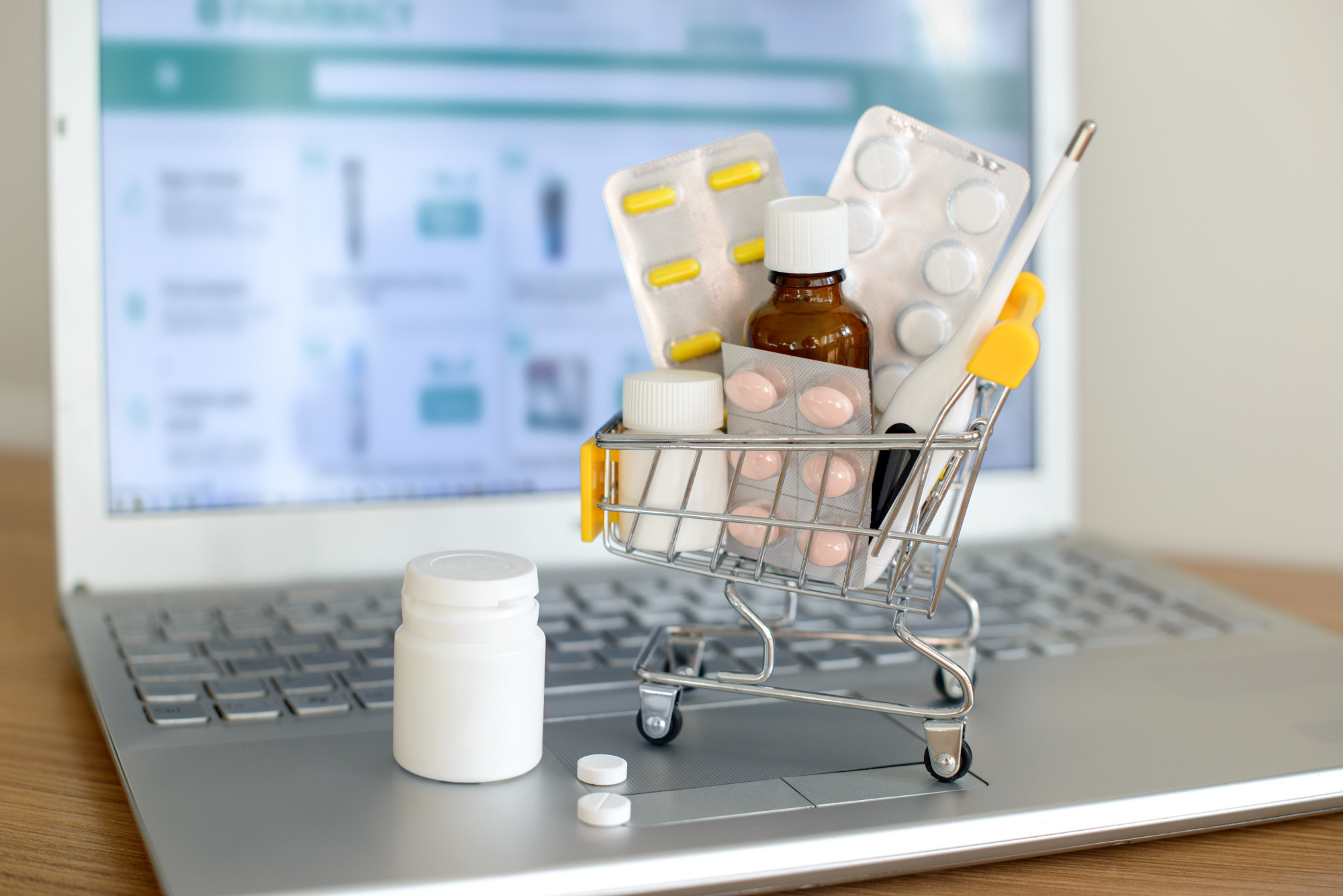 Shopping cart toy with medicaments in front of laptop screen with pharmacy web site on it. Pills, blister packs, medical bottles, thermometer set. Health care and internet shopping.