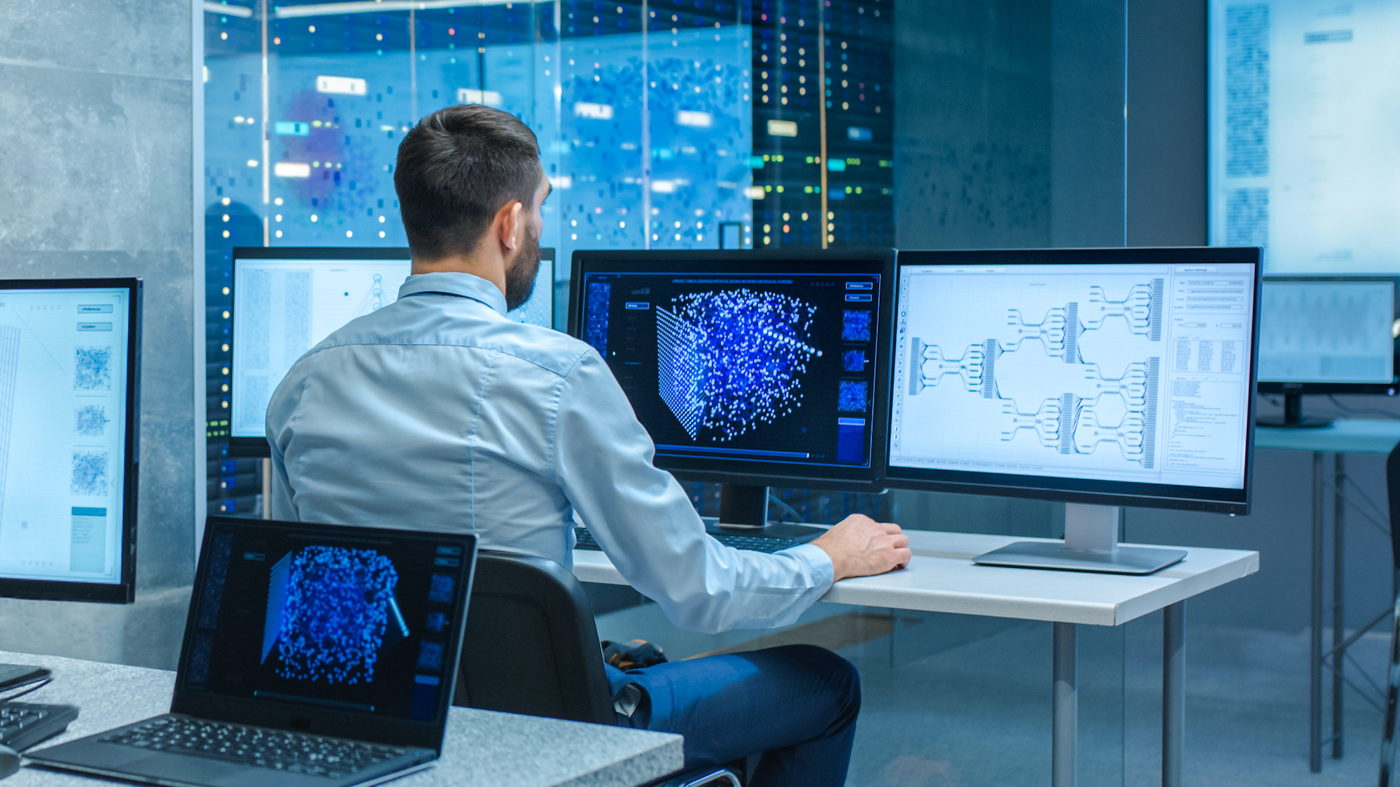 Beautiful Male Computer Engineer and Scientists Create Neural Network at His Workstation. Office is Full of Displays Showing 3D Representations of Neural Networks.